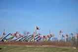 An array of boom lifts is lined up side by side on a grassy field, their arms extended upwards against a clear blue sky. The machines are in various colors including orange, yellow, and blue.