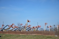 An array of boom lifts is lined up side by side on a grassy field, their arms extended upwards against a clear blue sky. The machines are in various colors including orange, yellow, and blue.