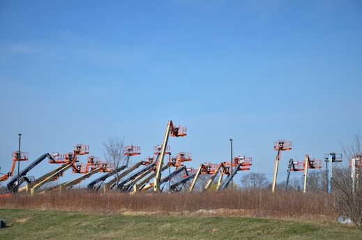 An array of boom lifts is lined up side by side on a grassy field, their arms extended upwards against a clear blue sky. The machines are in various colors including orange, yellow, and blue.