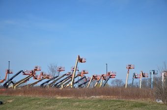 An array of boom lifts is lined up side by side on a grassy field, their arms extended upwards against a clear blue sky. The machines are in various colors including orange, yellow, and blue.