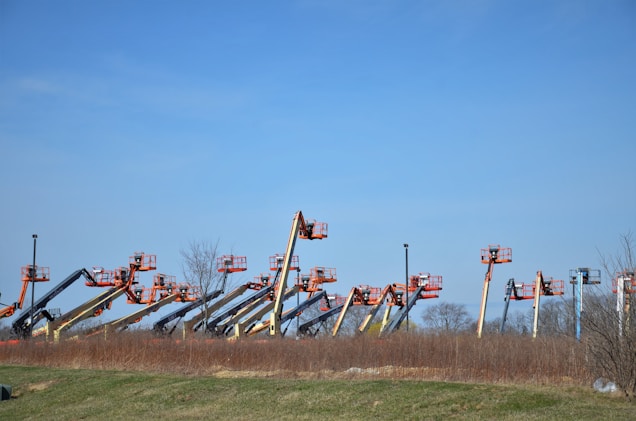 An array of boom lifts is lined up side by side on a grassy field, their arms extended upwards against a clear blue sky. The machines are in various colors including orange, yellow, and blue.