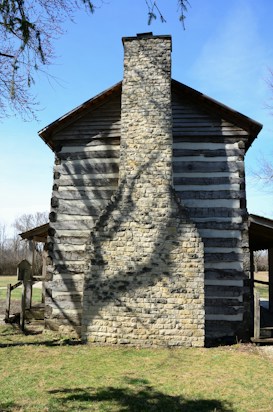 A rustic log cabin with a prominent stone chimney stands in a grassy area. Sunlight casts the shadow of a leafless tree on the chimney, creating an intricate pattern. The wooden structure has alternating dark and light logs, giving it a horizontally striped appearance. The surroundings include bare trees and clear blue sky in the background.