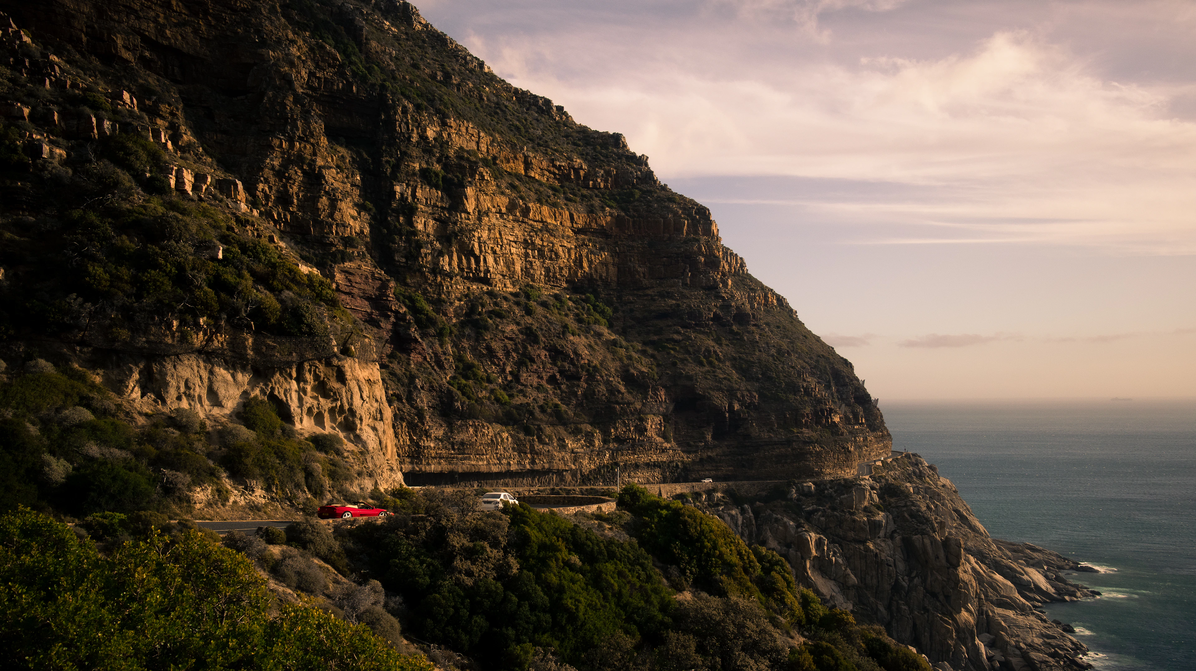 Cars on the narrow ledge of Chapman's Peak Drive, cut into a layered sandstone cliff above the Atlantic, in warm late afternoon light