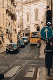 A bustling urban street scene featuring a tram, bus, and several cars moving along a narrow road lined with historic buildings. The cobblestone pavement has railway tracks embedded into it, and there are several pedestrians on the sidewalks. A traffic light signals green, and a road sign indicates a forward direction.