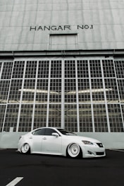 A sleek, white modified sedan with lowered suspension is parked in front of a large industrial hangar door labeled 'HANGAR NO1'. The hangar has a grid of windows and a corrugated metal facade, creating a backdrop that emphasizes the car's clean lines and customized features.