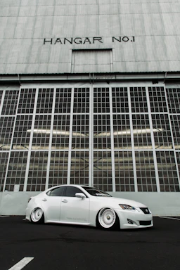 A sleek, white modified sedan with lowered suspension is parked in front of a large industrial hangar door labeled 'HANGAR NO1'. The hangar has a grid of windows and a corrugated metal facade, creating a backdrop that emphasizes the car's clean lines and customized features.