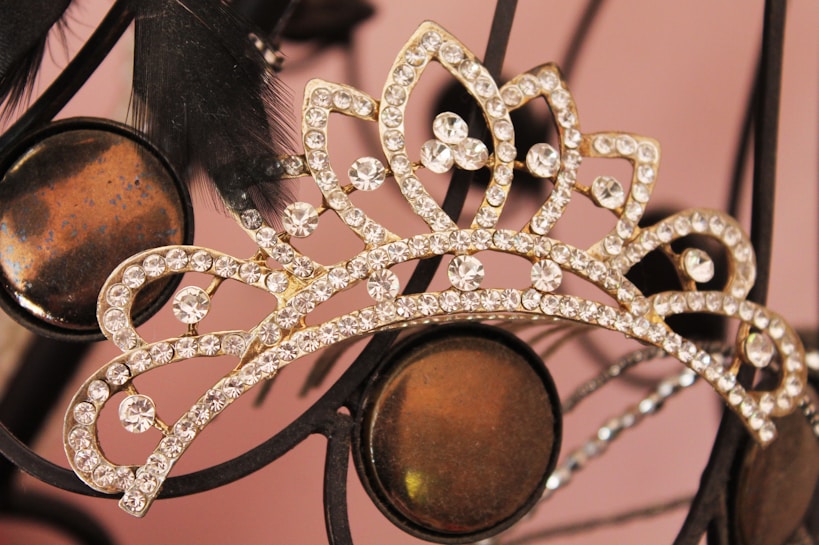 A jeweled tiara adorned with sparkling rhinestones is set against a soft focus background. The intricate design features a series of petal-like shapes, with each segment encrusted in small, clear stones. Dark feathers create an elegant contrast to the shimmering tiara, and circular metallic elements add a subtle touch of reflective color.