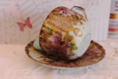 A decorative porcelain teacup and saucer featuring ornate golden patterns and floral motifs. The background includes a pale pink butterfly and faint text, providing a vintage and delicate aesthetic.