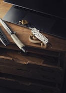 A rustic wooden desk displays a sleek knife with a wrapped grip, a multi-tool with a textured handle, part of a laptop, and a coiled rope. The arrangement suggests themes of adventure or outdoor activities.
