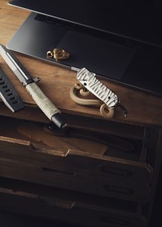 A rustic wooden desk displays a sleek knife with a wrapped grip, a multi-tool with a textured handle, part of a laptop, and a coiled rope. The arrangement suggests themes of adventure or outdoor activities.