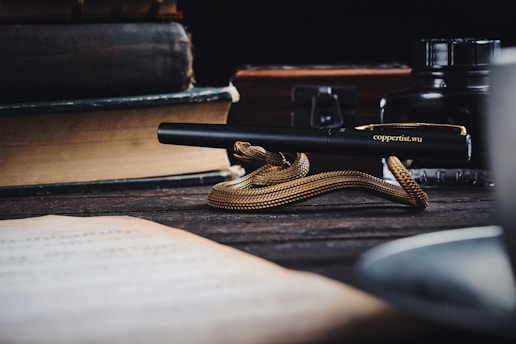 A golden snake-shaped pen lies elegantly on a dark wooden table, surrounded by old books and an ink bottle. The scene is composed with a vintage style, evoking a sense of timelessness and sophistication.