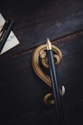 A sleek black fountain pen with a gold nib is placed horizontally on a dark, rustic wooden surface. Underneath the pen, there is a detailed golden serpent sculpture curled in an elegant spiral. To the left, part of a paper with handwritten text and the pen's cap are visible.