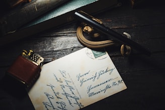 A vintage brown lighter and a fountain pen rest on top of a handwritten letter or postcard. The background consists of a dark wooden surface with a teal-colored book partially visible. A decorative rope or snake-like object is coiled around the pen.
