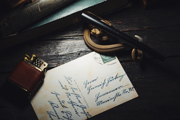 A vintage brown lighter and a fountain pen rest on top of a handwritten letter or postcard. The background consists of a dark wooden surface with a teal-colored book partially visible. A decorative rope or snake-like object is coiled around the pen.