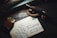A vintage brown lighter and a fountain pen rest on top of a handwritten letter or postcard. The background consists of a dark wooden surface with a teal-colored book partially visible. A decorative rope or snake-like object is coiled around the pen.