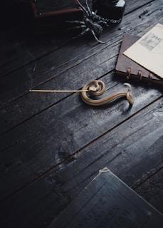 A dark, rustic wooden table holds a coil of rope, an incense stick sending a thread of smoke into the air, a stack of envelopes bound by a leather strap, and a metallic spider decoration. The lighting casts deep shadows and highlights, adding to the moody atmosphere.