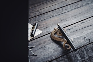 A black fountain pen rests on a rustic wooden surface, accompanied by a coil of a small golden snake figurine. Nearby, a piece of paper with handwritten text, partially obscured, lies alongside an uncapped black pen with a gold clip.