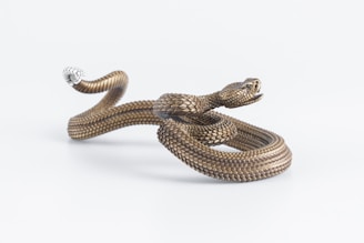 A coiled, bronze-colored snake-shaped object with detailed scales and a realistic, open-mouthed head. The snake appears to be artfully crafted, suggesting a decorative or artistic purpose. The white background accentuates the intricate detailing of the piece.