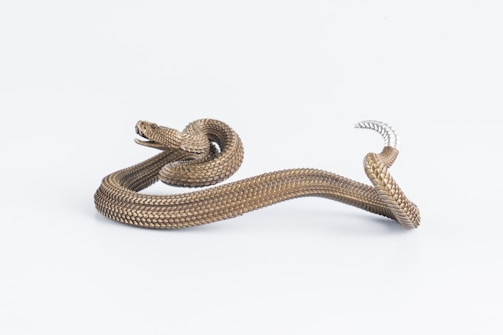 A snake with intricate, shiny scales and a strikingly realistic texture is set against a plain white background. Its body is partially coiled, displaying a dramatic curve and twist, with an open mouth showing fine detail.