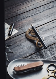 A fountain pen rests atop a coiled snake-shaped pen holder on a rustic wooden surface. Nearby, a piece of paper with cursive handwriting and the pen cap are partially visible. In the foreground, a tray holds a multi-tool knife and a pair of scissors, creating a vintage and artistic arrangement.