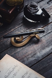 A sophisticated setting featuring a black fountain pen with gold accents rests atop a spiral, snake-shaped ornament on a wooden table. An open ink bottle is nearby, adding to the vintage atmosphere. A sheet of music paper is partially visible in the foreground.
