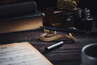A vintage setting featuring a fountain pen resting on a twisted golden rope, accompanied by an ink bottle on a wooden table. Nearby, a sheet of handwritten music is partially visible. The backdrop includes a stack of old books and a wooden box with a metal clasp, evoking a sense of nostalgia and creativity.