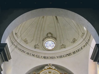 An ornate, domed ceiling featuring a circular stained glass window at its center. The architectural design includes intricate plasterwork and Latin inscriptions. The ceiling is painted in soft beige tones with decorative elements in light gold.