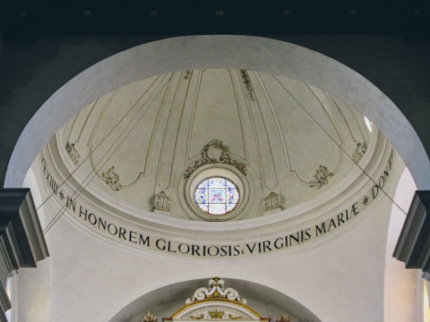 An ornate, domed ceiling featuring a circular stained glass window at its center. The architectural design includes intricate plasterwork and Latin inscriptions. The ceiling is painted in soft beige tones with decorative elements in light gold.