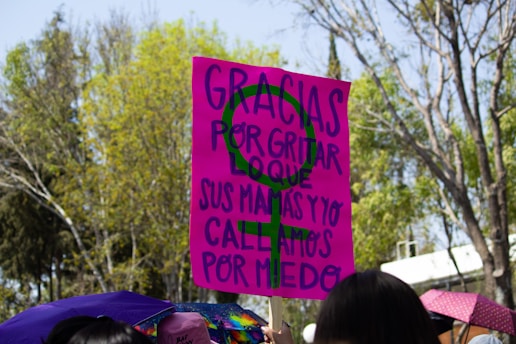 A bright pink protest sign with a green feminist symbol is held aloft, bearing the message in Spanish. The words express gratitude for speaking out despite fear. The sign is among a crowd, with scattered umbrellas in various colors visible below. Behind the scene, a backdrop of tall leafy trees under a clear sky suggests an outdoor setting.