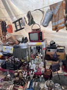 A collection of vintage and miscellaneous items displayed on a market stall. Objects include an old red television set, various decorative bottles and vases, a wall mirror, a pair of shoes, hanging fabrics, and assorted household items. A beaded necklace and other knick-knacks are visible, creating an eclectic and nostalgic atmosphere.