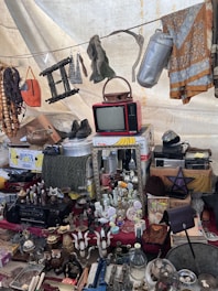 A collection of vintage and miscellaneous items displayed on a market stall. Objects include an old red television set, various decorative bottles and vases, a wall mirror, a pair of shoes, hanging fabrics, and assorted household items. A beaded necklace and other knick-knacks are visible, creating an eclectic and nostalgic atmosphere.