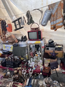 A collection of vintage and miscellaneous items displayed on a market stall. Objects include an old red television set, various decorative bottles and vases, a wall mirror, a pair of shoes, hanging fabrics, and assorted household items. A beaded necklace and other knick-knacks are visible, creating an eclectic and nostalgic atmosphere.