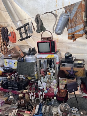 A collection of vintage and miscellaneous items displayed on a market stall. Objects include an old red television set, various decorative bottles and vases, a wall mirror, a pair of shoes, hanging fabrics, and assorted household items. A beaded necklace and other knick-knacks are visible, creating an eclectic and nostalgic atmosphere.