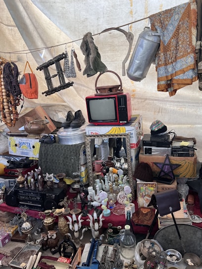 A collection of vintage and miscellaneous items displayed on a market stall. Objects include an old red television set, various decorative bottles and vases, a wall mirror, a pair of shoes, hanging fabrics, and assorted household items. A beaded necklace and other knick-knacks are visible, creating an eclectic and nostalgic atmosphere.