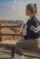 A woman sits on a wooden bench, overlooking a coastal cliff with a lighthouse in the distance. She holds a product in her hand, wearing sporty attire with a black jacket and brown leggings. The sky is partly cloudy, adding to a serene atmosphere.