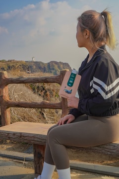 A woman sits on a wooden bench, overlooking a coastal cliff with a lighthouse in the distance. She holds a product in her hand, wearing sporty attire with a black jacket and brown leggings. The sky is partly cloudy, adding to a serene atmosphere.