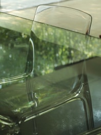 A transparent chair is placed in front of a glass table. The background shows an out-of-focus green foliage, likely seen through a glass window. The reflection on the glass surfaces enhances the scene's translucence and creates an interplay of light.