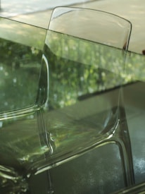 A transparent chair is placed in front of a glass table. The background shows an out-of-focus green foliage, likely seen through a glass window. The reflection on the glass surfaces enhances the scene's translucence and creates an interplay of light.