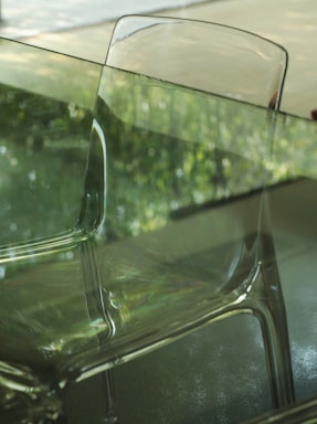 A transparent chair is placed in front of a glass table. The background shows an out-of-focus green foliage, likely seen through a glass window. The reflection on the glass surfaces enhances the scene's translucence and creates an interplay of light.