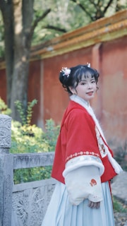 A woman in traditional attire stands beside an intricately carved stone railing, set against a backdrop of trees and a red wall. Her hair is styled with decorative ornaments, complementing her red and white outfit.