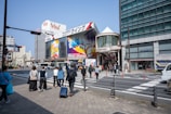 A busy urban street scene with pedestrians crossing a zebra crossing. The area is surrounded by modern buildings, including a large one with colorful advertisements and signage. People are dressed in casual clothing and some are carrying bags and pulling suitcases. The sky is clear and the lighting suggests it is daytime.