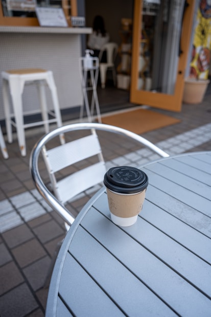 A takeaway coffee cup with a black lid sits on a round metal outdoor table. In the background, there's an open door to a cafe with some high stools and a few plants. The atmosphere seems casual and relaxed.