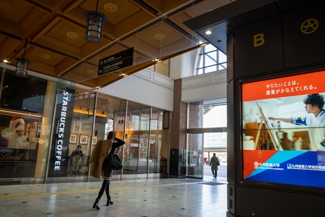 A modern indoor shopping area with a Starbucks Coffee shop and a digital advertisement display. People are walking by, and the building features a high ceiling with decorative elements and overhead lights. The advertisement display has text and an image of a person, likely promoting a service or product.