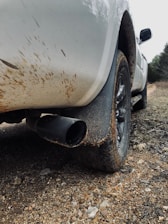 The image captures the rear section of a white vehicle with a focus on its tire and exhaust pipe. The car is parked on a rocky, dirt-covered surface, and the body of the vehicle shows signs of mud splatters, indicating it has been driven through a muddy area.