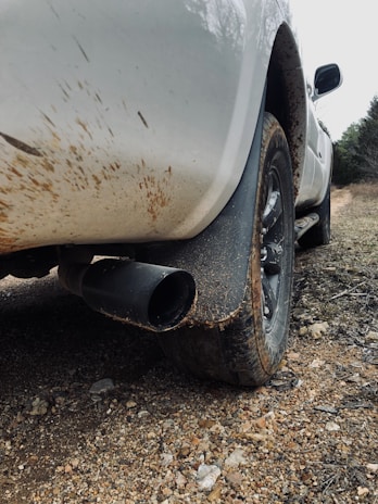 The image captures the rear section of a white vehicle with a focus on its tire and exhaust pipe. The car is parked on a rocky, dirt-covered surface, and the body of the vehicle shows signs of mud splatters, indicating it has been driven through a muddy area.
