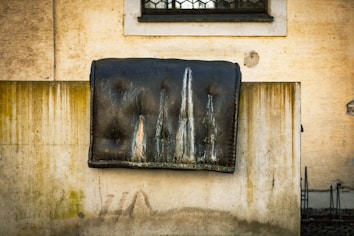 A worn and stained mattress placed over a concrete wall against an old, weathered building facade. The surface of the mattress is dark with prominent streaks and discolorations, while the wall shows signs of age and grime. The background features an old building with a small barred window.
