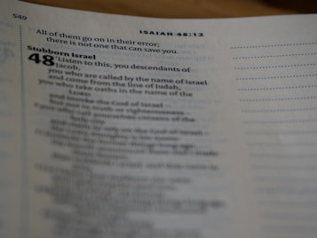 A close-up of a page from a religious text, specifically the book of Isaiah. The text is partially blurred, with a focus on a verse discussing 'Stubborn Israel.'