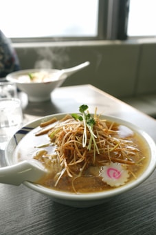 A steaming bowl of ramen with sliced pork, sprouts, and a decorative pink narutomaki sits on a table. In the background, another bowl of ramen is slightly out of focus, and the setting suggests a cozy dining environment with natural light coming through a window.