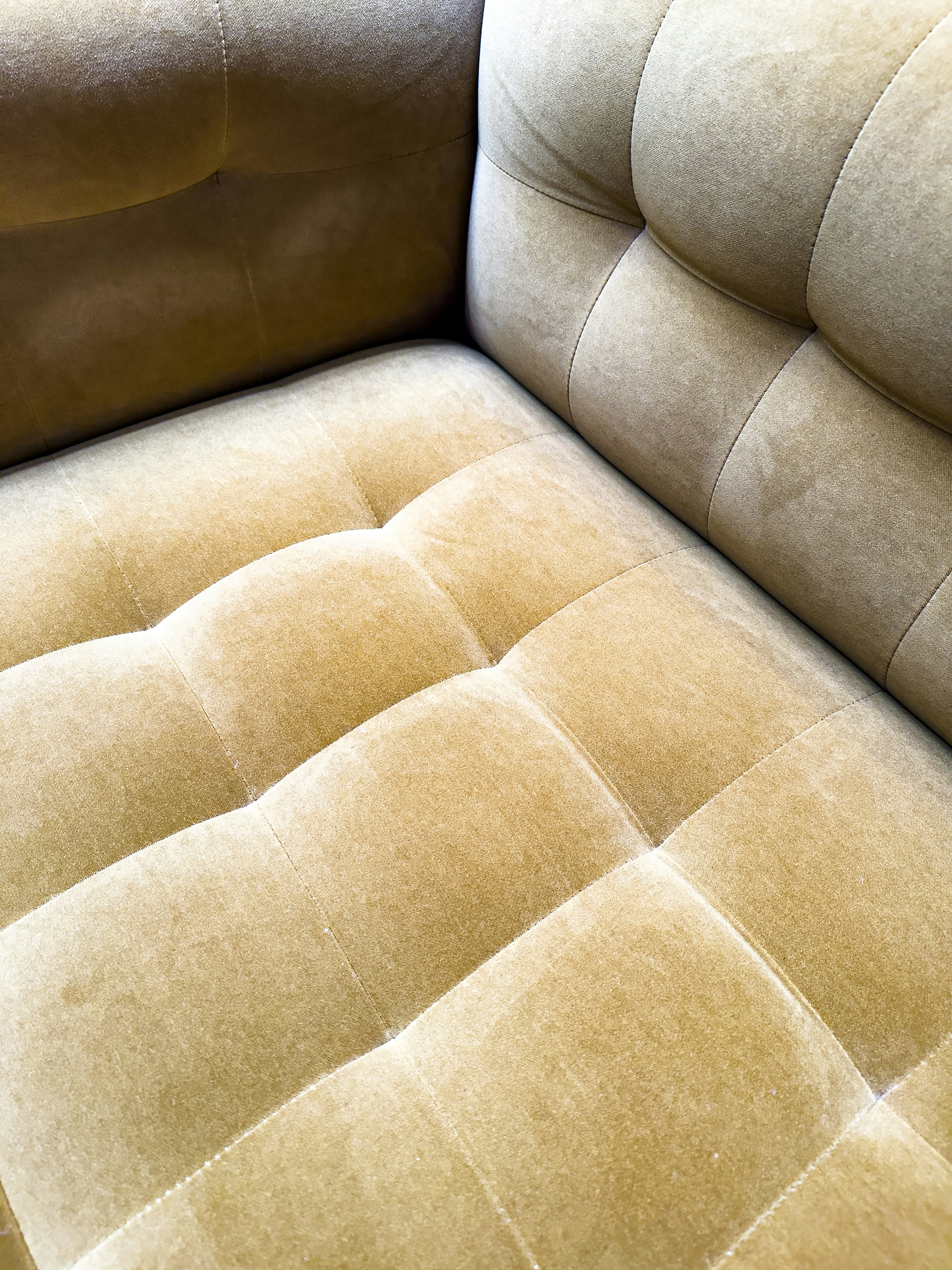 A close-up view of a plush, velvet-tufted cushion on a chair or sofa, showcasing its textured and luxurious finish in a muted golden brown color. The upholstery features a grid-like pattern with deep button tufting.