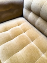 A close-up view of a plush, velvet-tufted cushion on a chair or sofa, showcasing its textured and luxurious finish in a muted golden brown color. The upholstery features a grid-like pattern with deep button tufting.